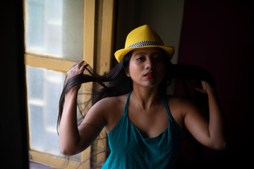 Portrait of an Asian/Japanese/Korean brunette young girl in greenish blue  western dress with yellow hat front of a window inside of a room. Fashion and cosplay photography.