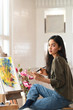 © Prathankarnpap - Photo of young beautiful artist holding a paint brush and drawing on painting canvas while sitting next to the arts accessories putting on small wooden table with studio windows as background.