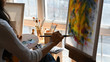 © Prathankarnpap - Cropped back image of young beautiful artist holding a brush and drawing on painting canvas while sitting next to the arts accessories putting on small wooden table with studio windows as background.