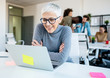 © Sanja - Woman looking at her work laptop while her colleagues discuss, joke and laugh.