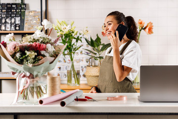 Wall Mural - Smiling female florist talking on mobile phone. Side view of woman working at flower shop.