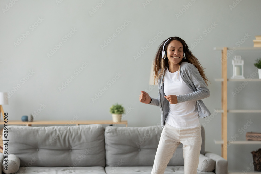 Happy millennial girl in modern headphones dance listen to music relax in living room, overjoyed young woman in earphones have fun entertain enjoy good quality sound at home, stress free concept - obrazy, fototapety, plakaty 