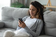 © fizkes - Smiling millennial Caucasian girl sit relax on cozy couch in living room browsing Internet on modern smartphone, happy young woman rest on comfortable sofa at home using cell texting or messaging