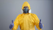 A man in yellow chemical protection suit and a respirator stands on a gray background and shows a thumbs up.