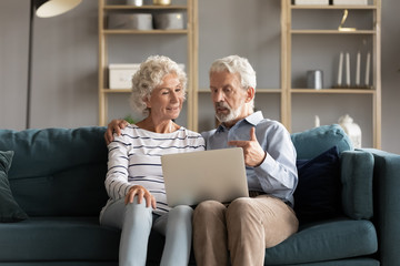 Wall Mural - Happy elderly husband and wife sit on couch in living room using modern computer together, smart old 60s middle-aged couple relax on comfortable sofa at home watch browse internet on laptop