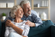 Smiling elderly husband and wife sit on sofa in living room look in distance dreaming and visualizing, happy optimistic mature 60s couple rest on couch at home think about happy future together