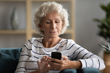 Modern Mature 50s Woman Sit On Couch In Living Room Texting Messaging On Smartphone, Smart Middle-aged Grandmother Relax At Home Using Browsing Internet On Cellphone, Elderly And Technology Concept