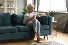 Calm Elderly Woman Sit On Comfortable Couch In Living Room Drink Tea Look In Window Distance Thinking Or Dreaming, Pensive Thoughtful Mature 50s Grandmother Enjoy Coffee Relax On Sofa At Home