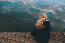 Young Girl With Long Blonde Hair Is Sitting On A Wooden Stage Looking In Clear Shallow Water With Reflecting Cloudy Sky In A Thoughtful Pose - Coming Of Age, Sadness And Sorrow Concept