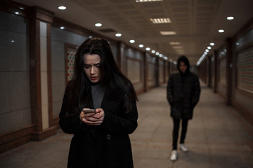 Wall Mural - a young beautiful woman walks through an underground passage at night, followed by a man in dark clothing with a hood on his head. a woman tries to dial the number of the rescue service on the phone