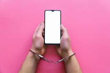 Men's hands in handcuffs hold a smartphone on a pink background. The concept of internet and gadget dependency.