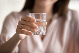 Fototapeta Łazienka - Cropped close up image focus on young woman reaching hand with glass of water to camera, proposing drinking fresh pure stilled liquid. Female nutritionist recommending daily rate, healthy habit.