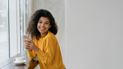 Beautiful African American woman using smartphone and mobile application for online shopping. Stylish hipster girl holding cellphone, communication, chatting, booking online, sitting in modern cafe