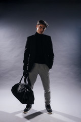 Canvas Print - Portrait of a young man with sharp jawline wearing a newsboy cap, holding a man bag while posing in studio on a white background.