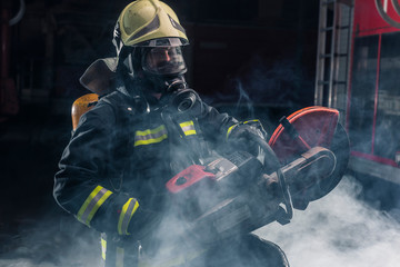 Portrait of a firefighter wearing full protective equipment posing with a chainsaw on his shoulder. Dark background with smoke and blue light.