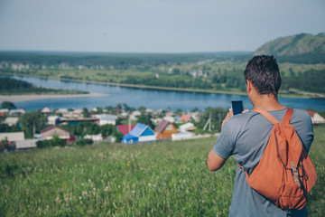 a man with a backpack photographs the horizon. A tourist travels all over the world. Village houses, forest and river in the background. The concept of summer, warmth, freedom, village life, sunburn