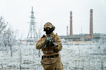 Wall Mural - Man in camouflage uniform hold machinegun. Soldier in the winter with factory on background. Horizontal photo side view.