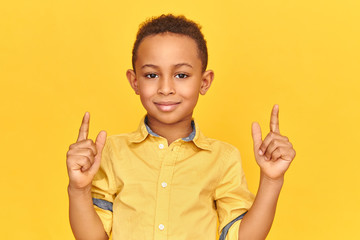 Handsome joyful Afro American boy having happy facial expression, smiling at camera, raising both index fingers, pointing upwards. Gestures, signs, symbols and non verbal communication concept