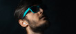 © Lalandrew - Close-up portrait of young hipster using wireless earphones, looking up wearing cyan sunglass, background of black color.