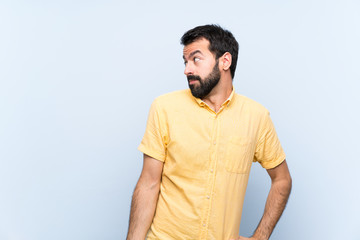 Wall Mural - Young man with beard over isolated blue background making doubts gesture looking side