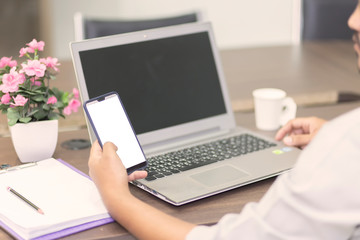 A man's hand is pressing on the smartphone screen on the table and the laptop is in the background. A business man eating coffee in a paper cup and pressing the phone with white screen.