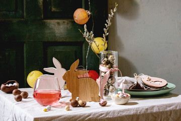 Easter table setting with wooden rabbit decoration, chocolate eggs and sweets, glasses of pink cocktail drinks, empty plates on white tablecloth. Lighting garlands. Holiday dinner.