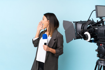 Reporter woman holding a microphone and reporting news over isolated blue background shouting with mouth wide open