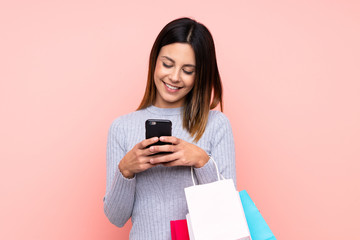 woman over isolated pink background holding shopping bags and writing a message with her cell phone 