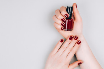 Beautiful dark red manicure with a bottle of nail polish in hands on a grey background. Procedures concept. Flat lay style.