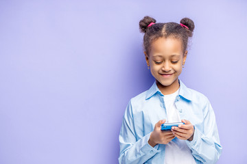 positive   african child girl with mobile phone in hands, isolated over purple background. sweet child chatting with friend or watching something interesting on phone