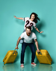 Couple in casual clothes. Girl sitting on back of her boyfriend, they smiling, guy holding two yellow suitcases. Blue background