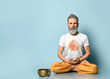 Yogi gray-haired man in dhoti clothes, holding rosary, sitting on floor in lotus pose on blue background. Singing bowl nearby