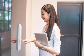 asian woman office assistance employer pointing and pressing on the button at the elevator while holding a tablet device with both hands on her stomach, wearing a long sleeves shirt and a pink skirt.