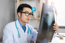 Male Asian Medical Doctor Looking And Examining A Bone Structure Of An X-ray Filter, Holding The Filter And Diagnosing His Patient Health, Wearing A Lab Coat Stethoscope Working In A Hospital Office
