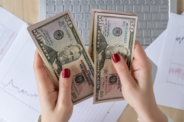 Time lapse. Woman freelancer holding banknotes fifty U.S. dollars in hand and counting profit, income. laptop with graphics and charts printed on the paper on office table. Flat lay