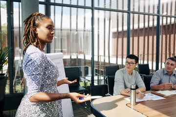 Pretty Hispanic businesswoman talking in front of colleagues in meeting room and explaining strategy of company development