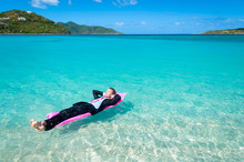 Relaxed Man In Suit Floating On A Bright Pink Pool Raft In Tropical Blue Waters