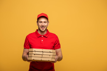 happy delivery man in red uniform holding pizza boxes isolated on yellow