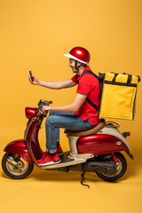 Wall Mural - side view of delivery man with backpack on scooter taking selfie on smartphone on yellow background