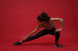 © ViDi Studio - Strong young african american sports fitness woman in sportswear posing working out isolated on red wall background studio portrait. Sport exercises healthy lifestyle concept. Stretching her legs.