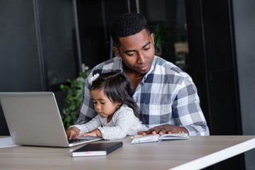 Young African-american father a designer at a remote work works at a computer sits at a table with his charming little two-year-old daughter. Concept of combining work and family