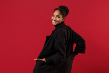 Back rear view of smiling young african american woman girl in black coat posing isolated on bright red wall background studio portrait. People lifestyle concept. Mock up copy space. Looking camera.