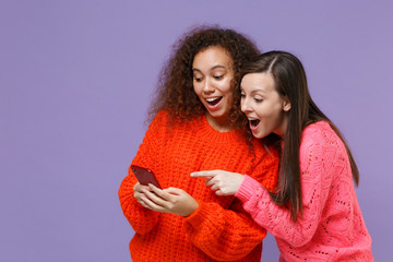 Surprised two young european african american women friends in knitted sweaters isolated on violet purple background. People lifestyle concept. Using point finger on mobile phone, typing sms message.