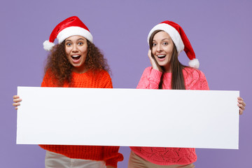 Wall Mural - Surprised european african american women friends in Christmas hat isolated on violet purple background. Happy New Year holiday concept. Mock up copy space. Hold blank sign board with place for text.