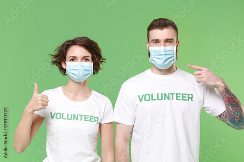 Two young friends couple in volunteer t-shirt isolated on pastel green background. Voluntary free work assistance help charity grace teamwork. Point index finger on sterile face mask showing thumb up.