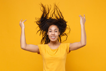 Funny young african american woman girl in casual t-shirt posing isolated on yellow orange background. People lifestyle concept. Mock up copy space. Have fun, throwing fluttering hair spreading hands.