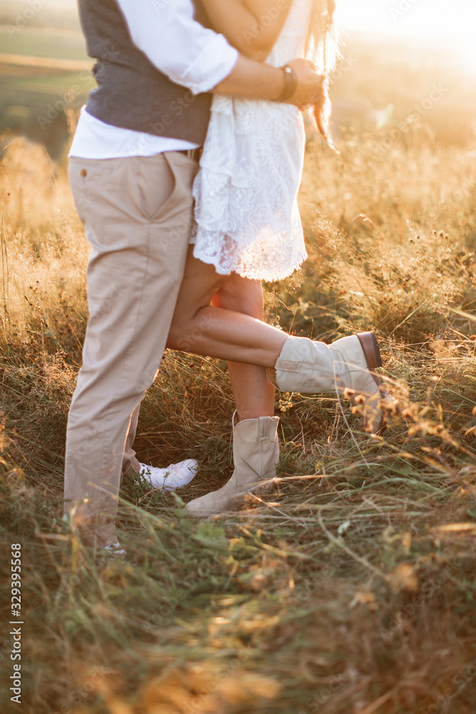Cropped image of legs of stylish rustic couple, boho woman in white dress  and cowboy boots and man in shirt and pants posing in sunny field, hugging  each other Stock Photo |