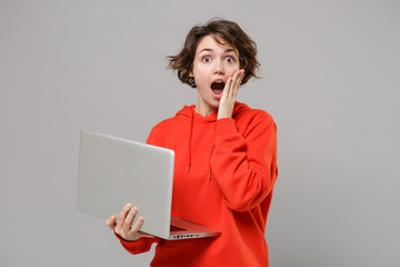 Canvas Print - Shocked young brunette woman girl in casual red hoodie posing isolated on grey background studio portrait. People lifestyle concept. Mock up copy space. Work on laptop pc computer put hand on cheek.