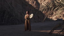 Young Woman In Dress Playing Tambourine In Mountains Shade In Desert At Sunset .