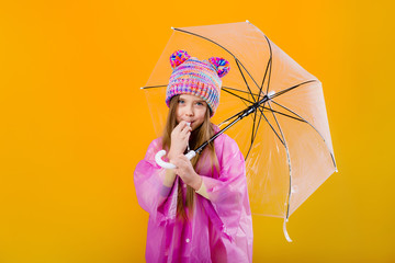 little girl in a pink raincoat and a knitted hat is holding an umbrella on a yellow background